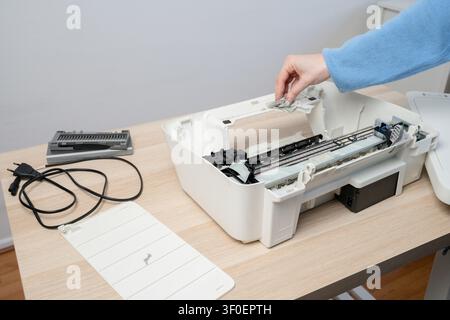A person wipes the interior of a disassembled white inkjet printer on a wooden desk, with removed parts, power cable and ink tank visible, illustratin Stock Photo