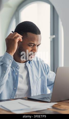 Young Black man concentrating on his laptop while working from a home office, surrounded by paperwork and natural light; thoughtful, productive, and e Stock Photo