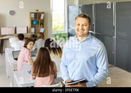 Confident teacher portrait in classroom with students at desks using laptops Stock Photo