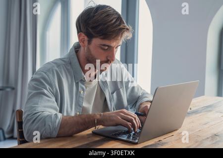 A focused young man types on a laptop at a wooden table in a bright home office, concentrating on remote work, freelance projects and online communica Stock Photo