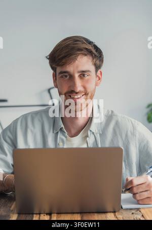 Smiling young man sits at a laptop in a bright home office, taking notes and working remotely with focus and relaxed confidence. Casual workspace conv Stock Photo