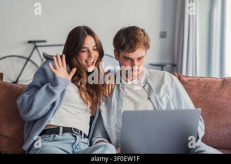 Happy young couple sitting on a sofa at home, looking at a laptop and waving during a video call; relaxed, engaged and sharing a joyful moment in a br Stock Photo