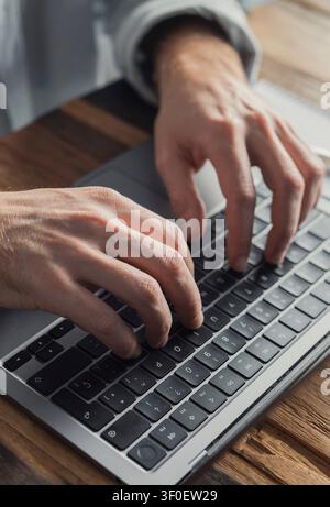 Close-up of a manís hands typing on a laptop keyboard on a wooden desk, suggesting remote work, writing, coding and productivity in a home office or m Stock Photo