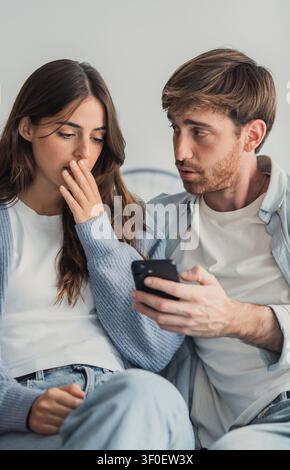 A young couple sits on a sofa as the man shows a smartphone to the woman; both look surprised and concerned, conveying tension, mistrust and emotional Stock Photo
