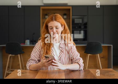 A young redhead woman sits at a wooden kitchen table in a modern home, smiling as she uses her smartphone. Natural light, casual striped shirt and rel Stock Photo