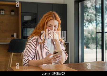A redheaded woman covers her mouth in surprised laughter as she reads a message on her smartphone, seated at a wooden table in a bright modern kitchen Stock Photo