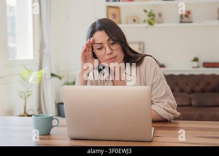 A young woman wearing glasses works on a laptop in a cozy living room, appearing thoughtful and focused while doing remote work or studying with a mug Stock Photo