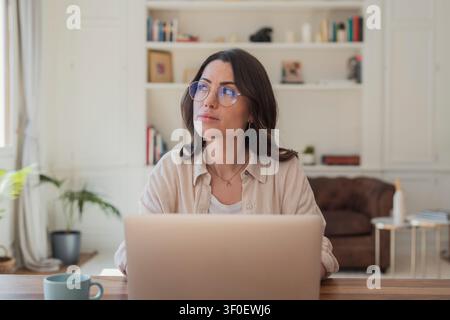 A thoughtful woman wearing glasses works on a laptop in a bright home office, pausing to look up as she thinks. Cozy workspace with bookshelf, sofa, c Stock Photo
