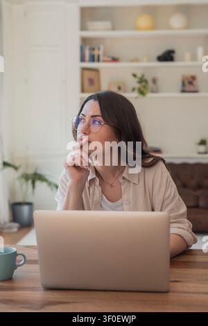 Young woman wearing glasses works on a laptop in a tidy home office, looking thoughtful as she plans and researches remote projects. Natural light, co Stock Photo