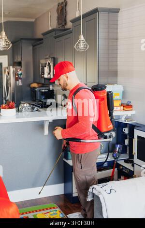 Man in red cap sprays kitchen with professional equipment. He wears backpack sprayer and focused expression. Bright lighting shows clean, modern kitch Stock Photo