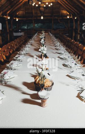 Long table set for formal dinner under warm lights. Wooden chairs flank both sides, ready for guests. Centerpiece holds bread, herbs, and rustic decor Stock Photo