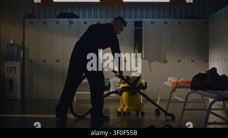 Professional Male Work in Messy Team Locker Room to Restore Order, Vacuums Tiled Floor Around Benches Scattered With Left Behind Sports Gear, Towels, Plastic Bottles, Working Diligently Clean Space. Stock Photo