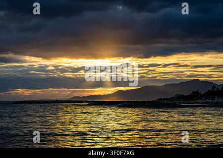 Dramatic Sunset Over Coastal Sea With Sun Rays Breaking Through Clouds And Mountain Silhouette, Province of Tarragona Stock Photo