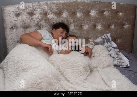 In a cozy bedroom setting, a grandmother lovingly watches her grandchild. The baby is lying in bed using a mobile phone, snug under a plush white blan Stock Photo