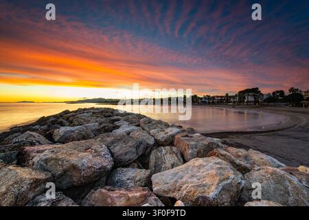 Glorious sunset paints the sky with vibrant orange and purple hues above a rocky jetty and tranquil beachfront, capturing a peaceful coastal seascape Stock Photo