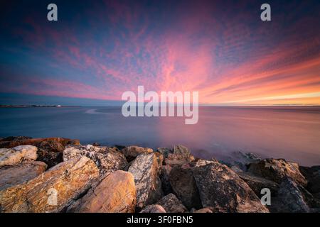Dramatic Pink Sunset Over Calm Ocean Seascape With Rocky Shoreline And Expansive Cloudscape, Cambrils, Catalonia, Spain Stock Photo