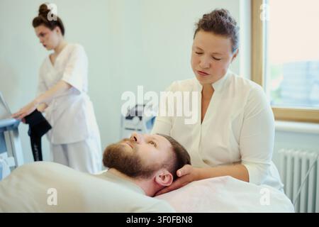 Young adult Caucasian woman therapist massaging neck of bearded Caucasian man during rehabilitation session while female colleague checking monitor. For wellness clinic marketing Stock Photo