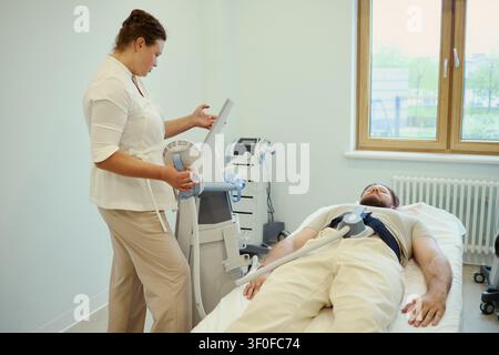 Wide shot of middle aged Caucasian woman operating medical device on middle aged Caucasian man lying on treatment bed. Ideal for healthcare therapy, rehab, wellness marketing Stock Photo