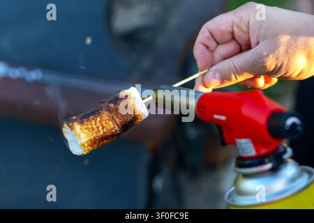 Toasting Marshmallow With Blowtorch on Skewer. Hand holds a skewer with a toasted marshmallow near a red blowtorch flame, showing browned sugar, smoke Stock Photo