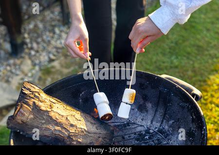 Marshmallows Toasting Over Outdoor Grill. Hands hold skewered marshmallows over an outdoor grill with ash, flame, firewood, grass, and warm sunlight. Stock Photo