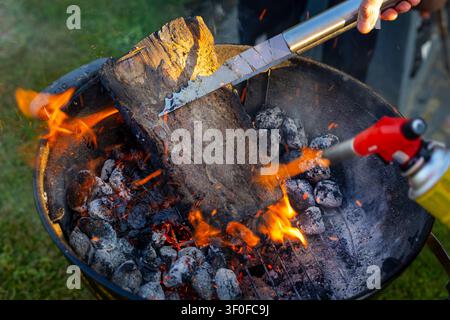 Firewood Ignited on Charcoal Grill With Blowtorch. Firewood burns on a charcoal grill as a blowtorch, tongs, flames, sparks, ash, and glowing coals Stock Photo