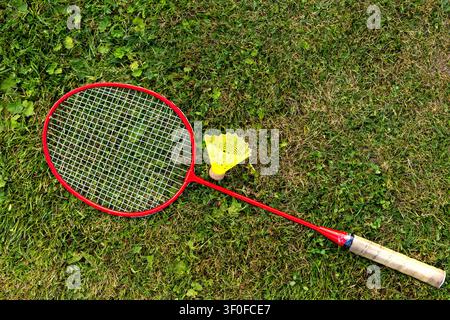 Badminton Racket and Shuttlecock on Grass. Red badminton racket and yellow shuttlecock lie on green grass in an outdoor top view scene. Stock Photo