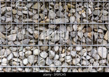 Gabion Stone Wall With Metal Wire Mesh. Close-up of a gabion wall filled with gray rocks and pebbles behind a metal wire grid. Stock Photo