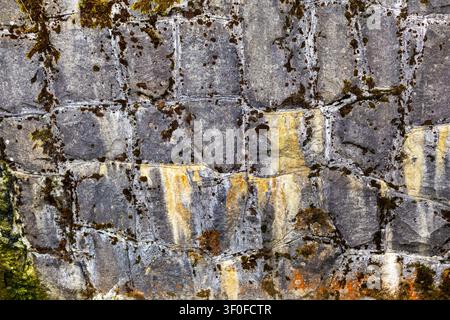 Mossy Stone Wall Texture With Mortar Lines. Mossy gray stone wall with dark slabs, pale mortar lines, yellow stains, rough texture, and weathered Stock Photo