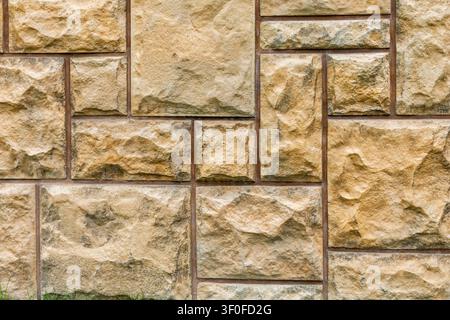 Beige Stone Veneer Wall Texture. Beige stone veneer blocks form a rough textured wall with brown grout lines and grass along the bottom edge. Stock Photo