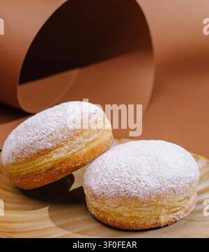 Two round donuts coated with powdered sugar sit on a wooden plate. The background features a smooth brown surface that adds warmth to the scene. The focus is on the sweet treats. Stock Photo