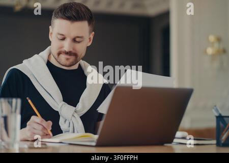 Young man reviews papers and writes notes beside laptop while working at desk indoors Stock Photo