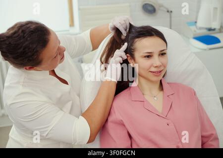 Middle aged female clinician examining young adult womans scalp and hair during consultation, checking skin condition. Useful for dermatology, trichology, wellness clinic marketing Stock Photo