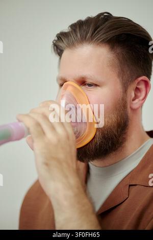 Vertical shot of Caucasian young adult man using inhalation mask for breathing treatment, managing respiratory care at home. Ideal for healthcare ads, asthma awareness, telehealth content Stock Photo