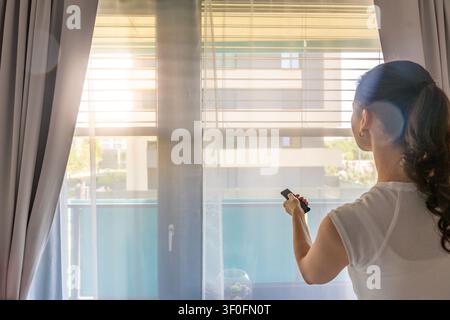 Woman using a remote control to adjust exterior window blinds in a modern apartment. Smart home comfort and energy efficient living concept. Stock Photo