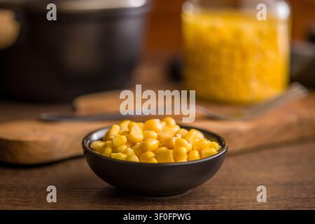 A black bowl of bright yellow sweet corn kernels rests on a rustic wooden table, with a blurred jar of canned corn and utensils behind it, ideal for f Stock Photo