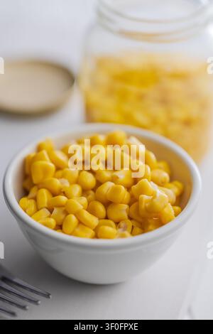 Close-up of bright yellow sweet corn kernels in a white bowl with a jar of corn in the background, suggesting fresh, simple home cooking, wholesome me Stock Photo