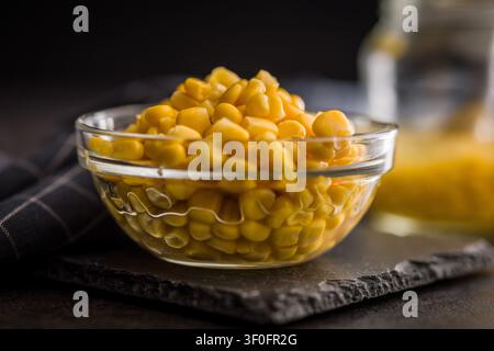 A glass bowl filled with glossy sweet corn kernels sits on a slate board against a dark, rustic background. Warm yellow tones highlight texture and fr Stock Photo