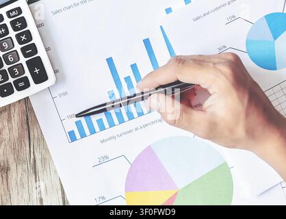 Hand holds pen above financial charts. Calculator sits on wooden desk. Person shows calm, focused work attitude. Great for finance or data analysis Stock Photo