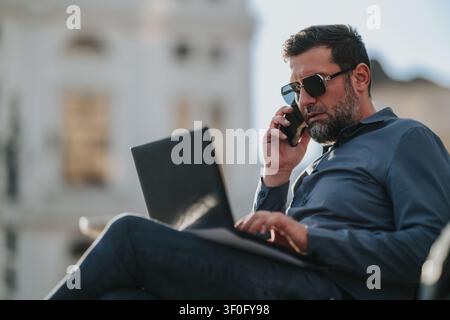 Businessman working remotely outdoors on laptop while talking on phone in casual attire Stock Photo