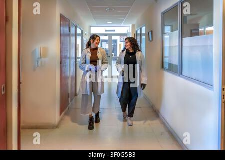 Two young female scientists in white lab coats and gloves smiling and talking while walking together through a bright institutional corridor with door Stock Photo