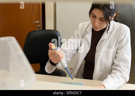 Young female scientist in white lab coat carefully using a micropipette at a desk in a bright laboratory, with a blurred foreground and office chair i Stock Photo