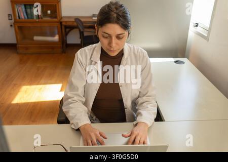 High angle view of a young female scientist in white lab coat typing on a laptop at a bright office desk, with a wooden bookcase and warm sunlight on Stock Photo