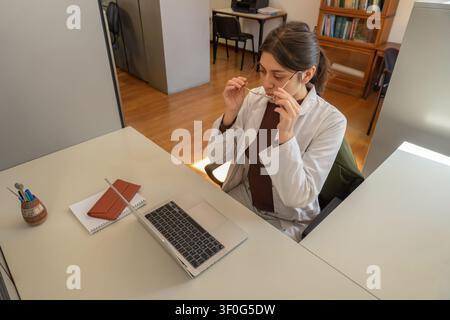 Young female scientist in white lab coat holding her glasses thoughtfully while sitting at a desk with a laptop, notebook and pen holder in a warm sun Stock Photo