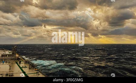 Horizontal dramatic shot from a ship deck showing magnificent golden sun rays breaking through dark storm clouds over a rough deep blue ocean with whi Stock Photo