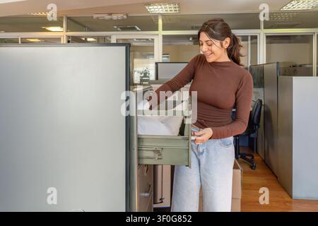 Young smiling brunette woman in casual clothes placing a white lab coat into an open drawer of a metal filing cabinet in a bright open plan office wit Stock Photo