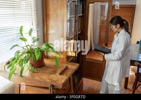 Young female scientist in white lab coat examining a large hardcover book just taken from an antique wooden bookcase in a bright office with indoor po Stock Photo