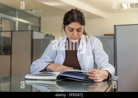 Young female scientist in white lab coat sitting at a glass table intently reading a large open book in a bright office with cubicle dividers and fili Stock Photo