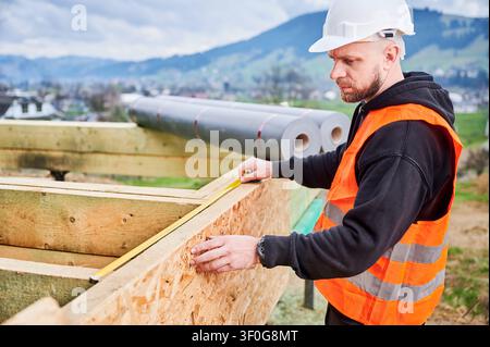 Construction worker in safety vest measuring wooden wall panel with tape, checking dimensions and alignment during house building process on outdoor site Stock Photo