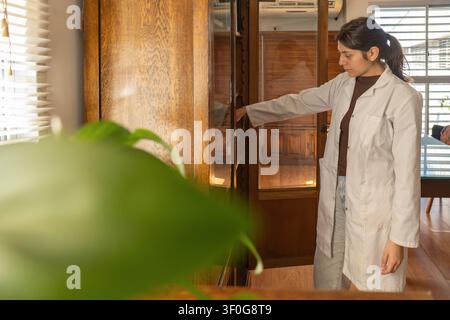 Creative shot through blurred plant foreground of a young female scientist in white lab coat closing an antique wooden bookcase with glass doors in a Stock Photo