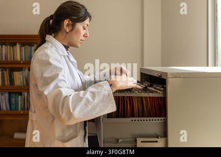 Young female scientist in white lab coat searching through hanging orange folders in an open filing cabinet drawer, with a bookcase of science books v Stock Photo
